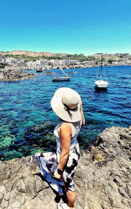 femme de dos avec un grand chapeau contemplant la mer debout sur les rochers a aci castello en sicile