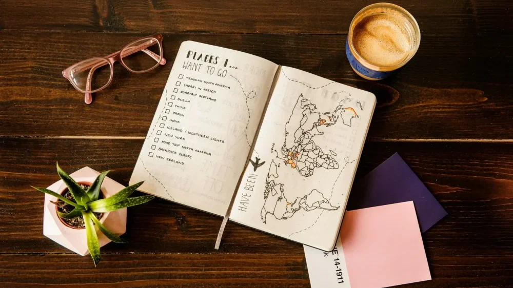 carnet ouvert sur une table bucket list voyage coché sur les pages. Café et lunettes posés a cote