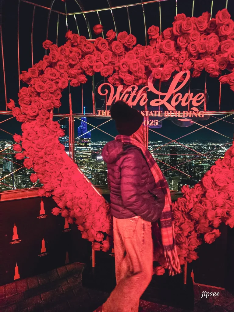 femme de dos en manteau et bonnet la nuit devant une decoration de coeur rouge en fleur siglé whith love empire state building