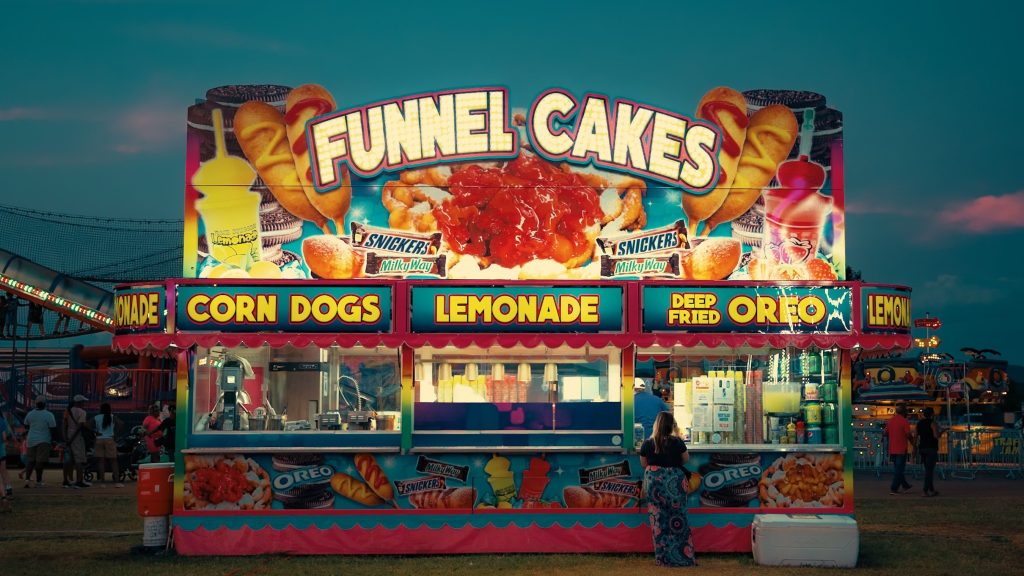 Colorful funnel cake stand illuminating a bustling carnival evening.