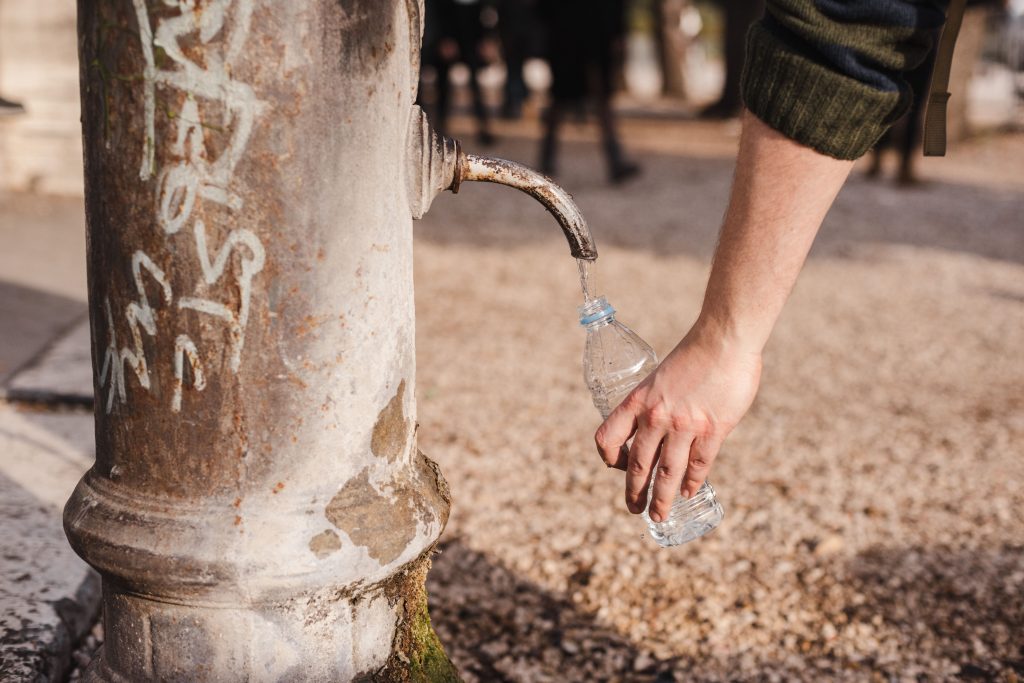 Crop unrecognizable person with bottle under old fountain tap with aqua flow on walkway in sunny town